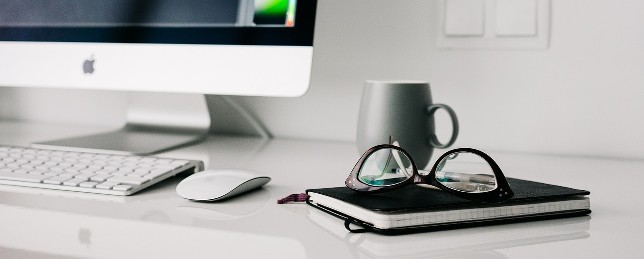 White office desk with a monitor, mouse, notebook, glasses, and a coffee mug